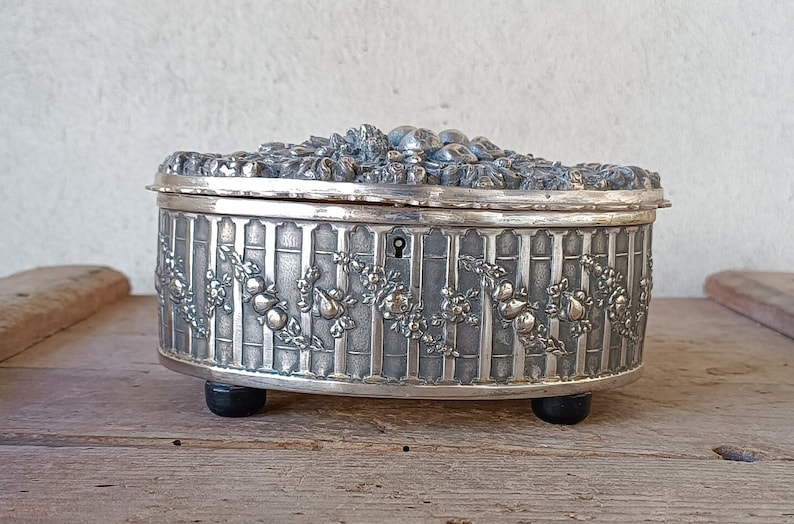 May include: A silver oval trinket box with an ornate floral design and a keyhole on the side. The box has three feet and is sitting on a wooden surface.