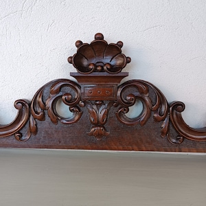 Könnte beinhalten: Ein dunkelbraunes Holzkopfteil mit aufwendigen Schnitzereien. Das Kopfteil hat ein kronenartiges Design mit einem Muschelmotiv und einem dekorativen Rankenmuster.