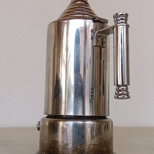 Peut inclure: Une cafeti&egrave;re vintage argent&eacute;e avec un dessus conique et une poign&eacute;e. La cafeti&egrave;re a un corps cylindrique et repose sur une base en bois fonc&eacute;. Le design est r&eacute;fl&eacute;chissant et semble &ecirc;tre en m&eacute;tal.