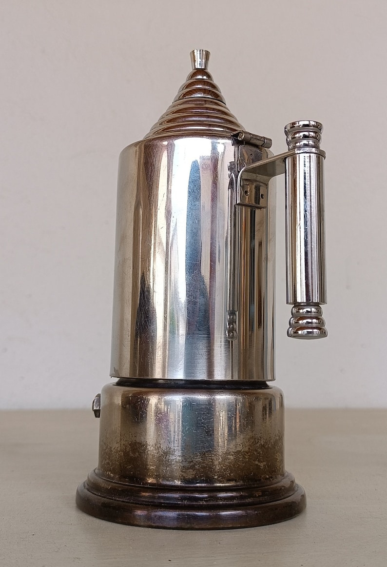 Peut inclure: Une cafeti&egrave;re vintage argent&eacute;e avec un dessus conique et une poign&eacute;e. La cafeti&egrave;re a un corps cylindrique et repose sur une base en bois fonc&eacute;. Le design est r&eacute;fl&eacute;chissant et semble &ecirc;tre en m&eacute;tal.