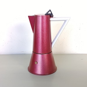 Pode incluir: Uma cafeteira de espresso vermelha e branca com uma pega triangular. A cafeteira tem um acabamento texturizado e metálico.