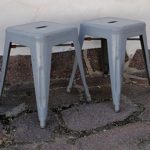 May include: Two grey metal stools with a simple design. The stools have a square seat and four legs.