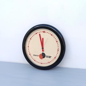 Peut inclure: Une horloge murale ronde avec un cadre noir et un cadran crème. L'horloge présente des aiguilles rouges et des points rouges marquant les heures. Les logos Pirelli et Partner System sont visibles en bas.