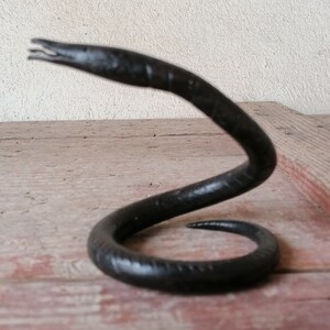 K&ouml;nnte beinhalten: Eine schwarze Schlangenskulptur aus Metall mit einem gewundenen K&ouml;rper und einem erhobenen Kopf. Die Skulptur steht auf einer Holzoberfl&auml;che.