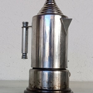 Peut inclure: Une cafeti&egrave;re vintage argent&eacute;e avec un dessus conique et un bec verseur triangulaire. La cafeti&egrave;re a un corps cylindrique et une poign&eacute;e. La base est marron fonc&eacute;. Le design rappelle les ustensiles de cuisine modernes du milieu du si&egrave;cle.