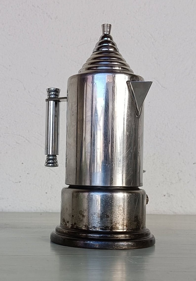 Peut inclure: Une cafeti&egrave;re vintage argent&eacute;e avec un dessus conique et un bec verseur triangulaire. La cafeti&egrave;re a un corps cylindrique et une poign&eacute;e. La base est marron fonc&eacute;. Le design rappelle les ustensiles de cuisine modernes du milieu du si&egrave;cle.