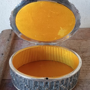 May include: A silver trinket box with an ornate design and a yellow velvet lining. The box is open and the interior is visible.