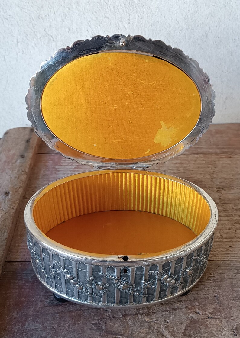 May include: A silver trinket box with an ornate design and a yellow velvet lining. The box is open and the interior is visible.