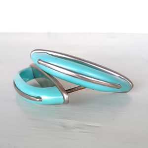 Könnte beinhalten: Zwei türkisblaue Schrankknöpfe mit silbernem Besatz. Die Knöpfe sind oval geformt und haben ein einzelnes Schraubenloch.