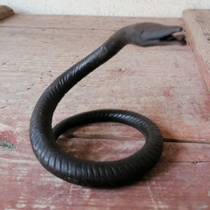 K&ouml;nnte beinhalten: Eine schwarze Schlangenskulptur aus Metall mit einem gewundenen K&ouml;rper und einem erhobenen Kopf. Die Skulptur hat eine strukturierte Oberfl&auml;che und besteht aus einem einzigen St&uuml;ck Metall.