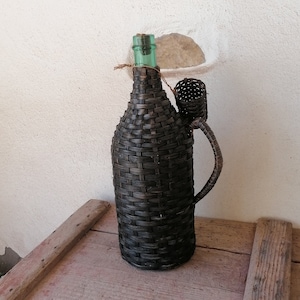 May include: A black wicker bottle with a green glass top and a wicker handle. The bottle is sitting on a wooden surface.