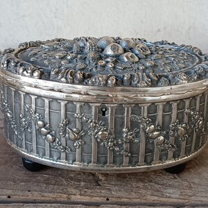 May include: A silver-toned trinket box with an ornate design featuring a fruit motif. The box has a hinged lid and a keyhole.