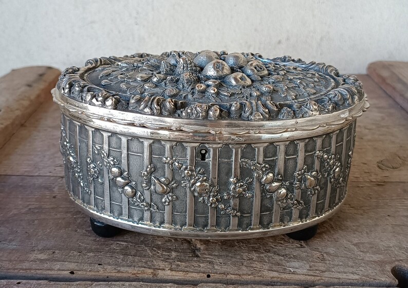 May include: A silver-toned trinket box with an ornate design featuring a fruit motif. The box has a hinged lid and a keyhole.