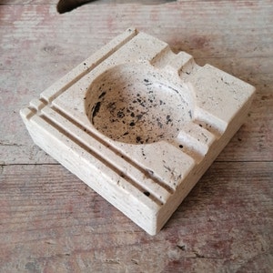 May include: A square, off-white, stone ashtray with a round, indented centre and three grooves around the edge.