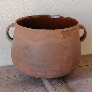 Puede incluir: Una olla grande de barro marrón con dos asas. La olla tiene una superficie rugosa y texturizada y parece estar hecha a mano.
