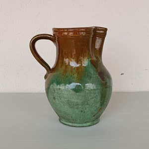 Puede incluir: Una jarra de cerámica esmaltada marrón y verde con asa. La jarra tiene una boca ancha y un cuerpo redondeado.