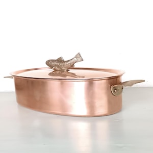 May include: An oval copper casserole dish with a fish-shaped lid handle. The dish has two handles on the sides and is made of polished copper.