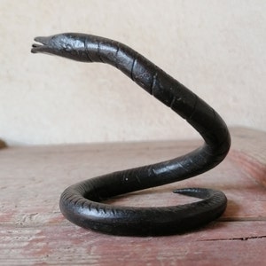 K&ouml;nnte beinhalten: Eine schwarze Metallsculptur einer Schlange in einer gewickelten Position. Die Schlange ist aus glattem, dunklem Metall gefertigt und hat ein realistisches Aussehen.