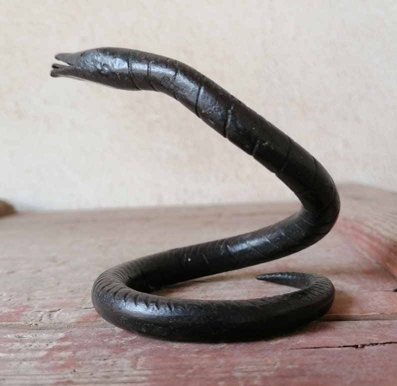 K&ouml;nnte beinhalten: Eine schwarze Metallsculptur einer Schlange in einer gewickelten Position. Die Schlange ist aus glattem, dunklem Metall gefertigt und hat ein realistisches Aussehen.
