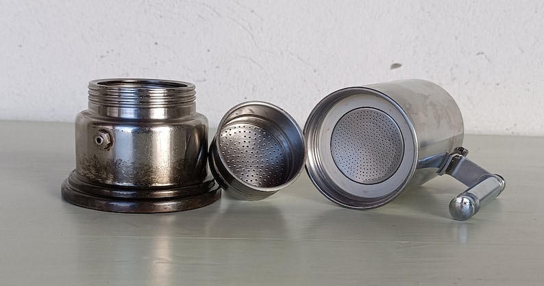 Peut inclure: Une cafeti&egrave;re italienne en acier inoxydable d&eacute;mont&eacute;e. Les composants comprennent la base, le panier-filtre et la chambre sup&eacute;rieure avec une poign&eacute;e. Les pi&egrave;ces sont dispos&eacute;es sur une surface claire, mettant en valeur l'&eacute;quipement de pr&eacute;paration du caf&eacute;.