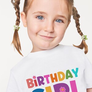Puede incluir: Una camiseta blanca con las palabras "Birthday Girl" impresas en letras de colores. Las letras est&aacute;n delineadas en negro y tienen un estilo juguet&oacute;n y caprichoso. Una peque&ntilde;a flor rosa con un centro amarillo est&aacute; al lado de la palabra "Girl".