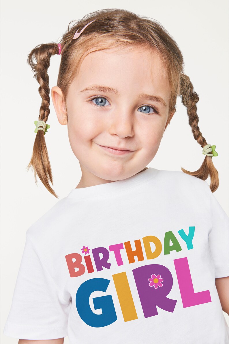 Puede incluir: Una camiseta blanca con las palabras "Birthday Girl" impresas en letras de colores. Las letras est&aacute;n delineadas en negro y tienen un estilo juguet&oacute;n y caprichoso. Una peque&ntilde;a flor rosa con un centro amarillo est&aacute; al lado de la palabra "Girl".