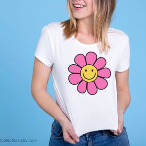 May include: White t-shirt with a pink flower graphic featuring a yellow smiley face.