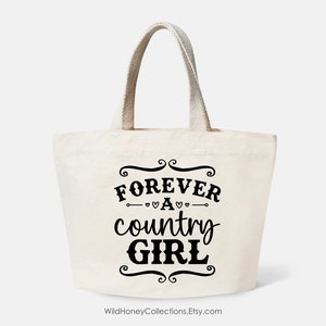 Puede incluir: Un bolso de tela de color crema con dos asas resistentes. La parte frontal presenta un texto en negro que dice FOREVER A country GIRL, enmarcado por adornos decorativos y peque&ntilde;os corazones encima y debajo de la palabra A.