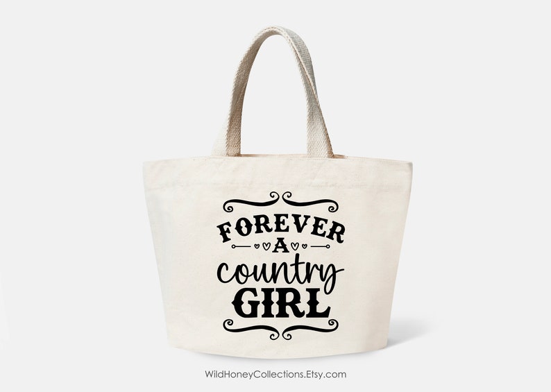 Puede incluir: Un bolso de tela de color crema con dos asas resistentes. La parte frontal presenta un texto en negro que dice FOREVER A country GIRL, enmarcado por adornos decorativos y peque&ntilde;os corazones encima y debajo de la palabra A.