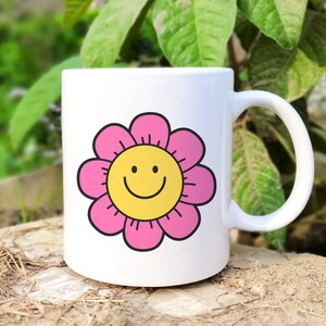 May include: White ceramic mug featuring a pink flower design with a yellow smiley face in the centre. The mug has a handle.