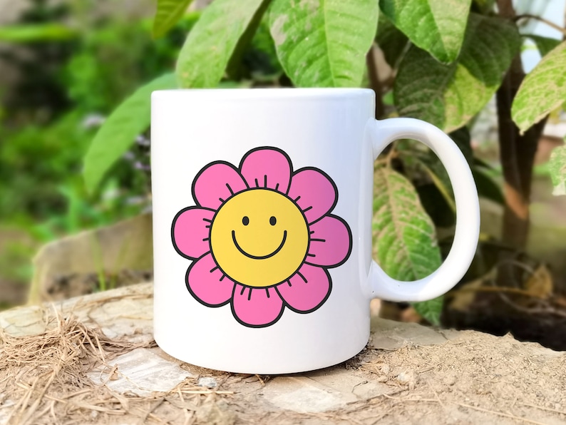 May include: White ceramic mug featuring a pink flower design with a yellow smiley face in the centre. The mug has a handle.