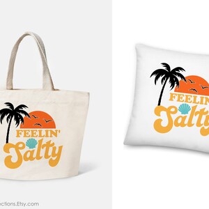 May include: A canvas tote bag and a white pillow with a beach-themed design. The design includes a black palm tree, an orange sunset, and the words "FEELIN' Salty" in orange and blue. The tote bag has beige handles.