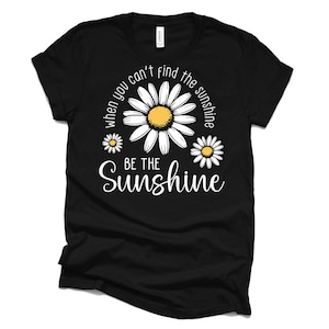 K&ouml;nnte beinhalten: Schwarzes T-Shirt mit wei&szlig;em Text, der lautet: "Wenn du die Sonne nicht finden kannst, sei die Sonne." Der Text ist von drei wei&szlig;en G&auml;nsebl&uuml;mchen mit gelben Zentren umgeben.