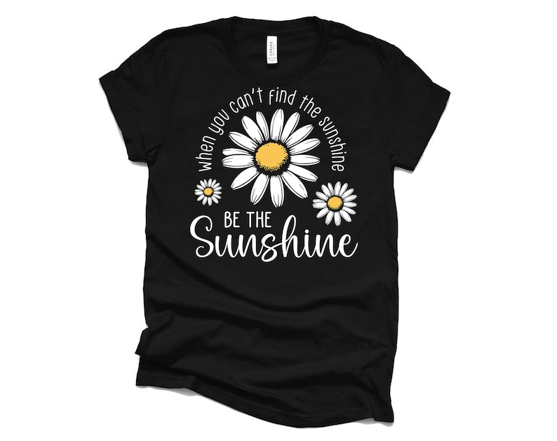 K&ouml;nnte beinhalten: Schwarzes T-Shirt mit wei&szlig;em Text, der lautet: "Wenn du die Sonne nicht finden kannst, sei die Sonne." Der Text ist von drei wei&szlig;en G&auml;nsebl&uuml;mchen mit gelben Zentren umgeben.
