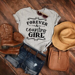 Puede incluir: Una camiseta gris jaspeado con el texto negro FOREVER A country GIRL, botas vaqueras de cuero marr&oacute;n, pantalones cortos de mezclilla azul oscuro con cintur&oacute;n de cuero marr&oacute;n, un sombrero de paja, un bolso bandolera de cuero marr&oacute;n y gafas de sol.