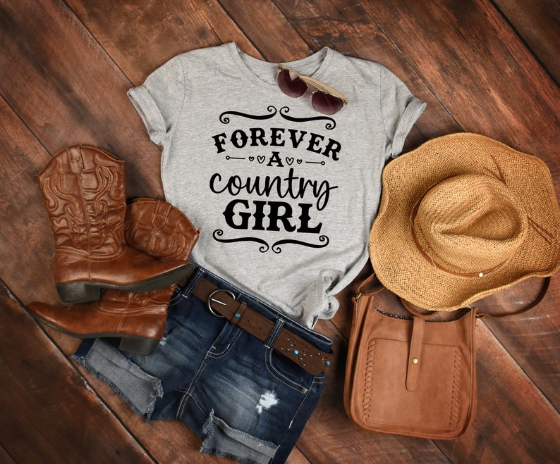 Puede incluir: Una camiseta gris jaspeado con el texto negro FOREVER A country GIRL, botas vaqueras de cuero marr&oacute;n, pantalones cortos de mezclilla azul oscuro con cintur&oacute;n de cuero marr&oacute;n, un sombrero de paja, un bolso bandolera de cuero marr&oacute;n y gafas de sol.