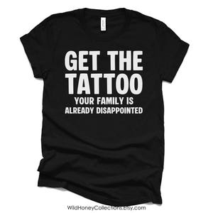 Op de afbeelding: Een zwart t-shirt met korte mouwen en witte tekst op de borst: GET THE TATTOO YOUR FAMILY IS ALREADY DISAPPOINTED. Het kledingstuk heeft een ronde hals en een standaard pasvorm, met een wit merklabel zichtbaar aan de binnenkant van de kraag.