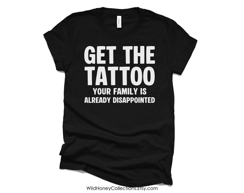 Op de afbeelding: Een zwart t-shirt met korte mouwen en witte tekst op de borst: GET THE TATTOO YOUR FAMILY IS ALREADY DISAPPOINTED. Het kledingstuk heeft een ronde hals en een standaard pasvorm, met een wit merklabel zichtbaar aan de binnenkant van de kraag.