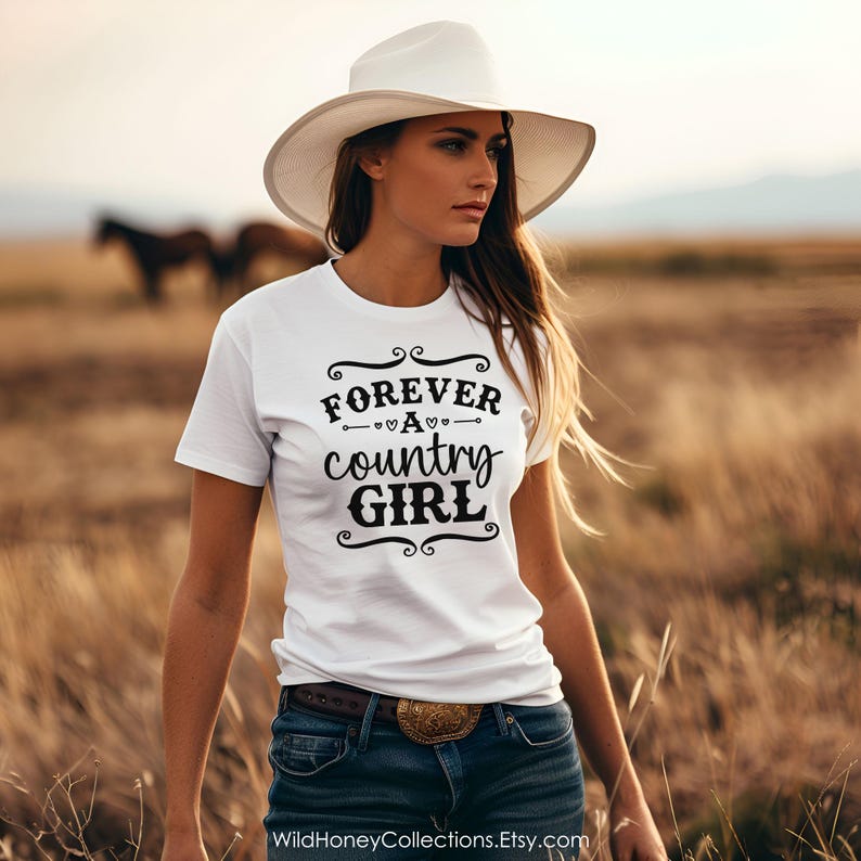 Puede incluir: Una camiseta blanca de manga corta con el texto en negro FOREVER A country GIRL, adornado con filigranas decorativas y peque&ntilde;os iconos de corazones. La prenda se combina con unos vaqueros azul oscuro y un sombrero blanco de ala ancha.
