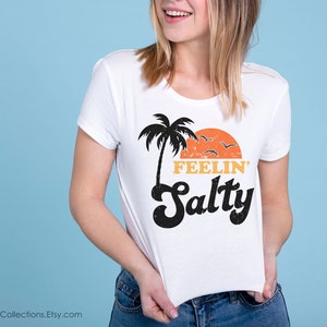 May include: White t-shirt with a retro design featuring a black palm tree, orange sunset, and the words "FEELIN' Salty" in black and orange. The shirt is being held up against a light blue background.