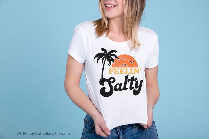 May include: White t-shirt with a retro design featuring a black palm tree, orange sunset, and the words "FEELIN' Salty" in black and orange. The shirt is being held up against a light blue background.
