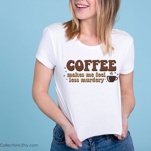 May include: White t-shirt with brown text that reads "COFFEE makes me feel less murdery."  A small brown coffee cup icon is next to the text.