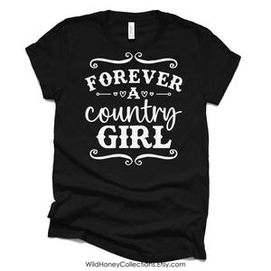Puede incluir: Una camiseta negra de manga corta con el texto impreso en blanco FOREVER A country GIRL, decorada con remolinos y peque&ntilde;os iconos de corazones centrados en el pecho de la prenda, que cuenta con un cuello redondo est&aacute;ndar.