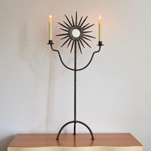 May include: A dark metal candelabra featuring a central sunburst design with a small circular mirror at its core. The structure has two curved arms holding lit cream-colored taper candles, supported by a tall vertical rod and a curved base.