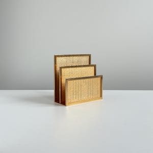May include: A tiered letter holder featuring three rectangular compartments with a woven cane texture and a metallic gold-toned frame. The structure is arranged in a stepped formation, allowing for the organized storage of documents or mail.