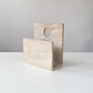 May include: A beige stone magazine holder featuring a rectangular base and a tall back panel with a circular cutout near the top. The natural material displays a textured, marbled surface with subtle variations in color and grain throughout the piece.