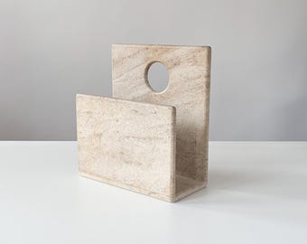 May include: A beige stone magazine holder featuring a rectangular base and a tall back panel with a circular cutout near the top. The natural material displays a textured, marbled surface with subtle variations in color and grain throughout the piece.