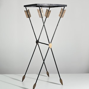 May include: A tall, slender accent table featuring a square black top supported by four thin, black metal legs designed to resemble crossed arrows. The arrow-shaped legs include gold-toned fletching at the top and gold-toned arrowheads at the base.