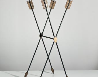 May include: A tall, slender accent table featuring a square black top supported by four thin, black metal legs designed to resemble crossed arrows. The arrow-shaped legs include gold-toned fletching at the top and gold-toned arrowheads at the base.