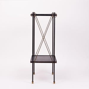May include: A black metal plant stand with a rectangular shelf and four legs. The stand features brass accents on the legs and decorative elements at the top. The stand has a minimalist design.